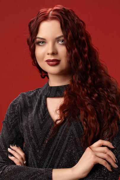 Beautiful sexy woman is posing in a knitted dress on a red background. Evening makeup. Long, healthy, red curly hair. - Photo, Image
