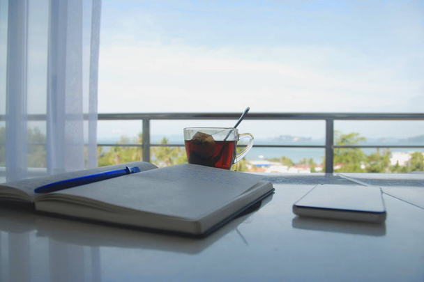 Otwórz Notatnik z długopisem, kieliszek herbaty i mobil telefon leżący na stole, z niesamowitym widokiem na morze w tle - Zdjęcie, obraz