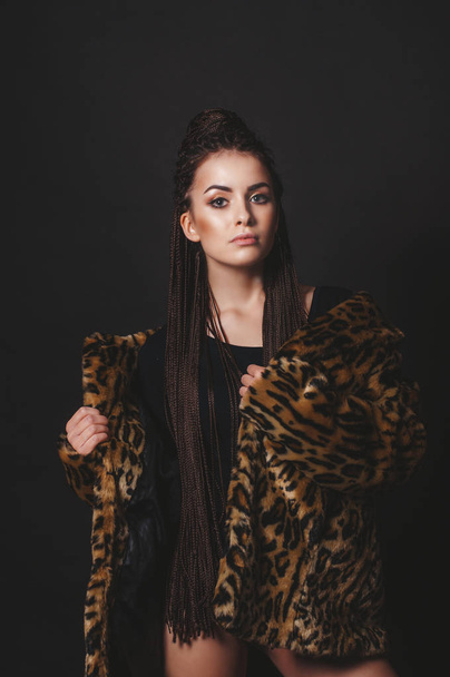 Young attractive girl with long afro-braids in a fur coat under leopard fur over black background - Foto, Imagem