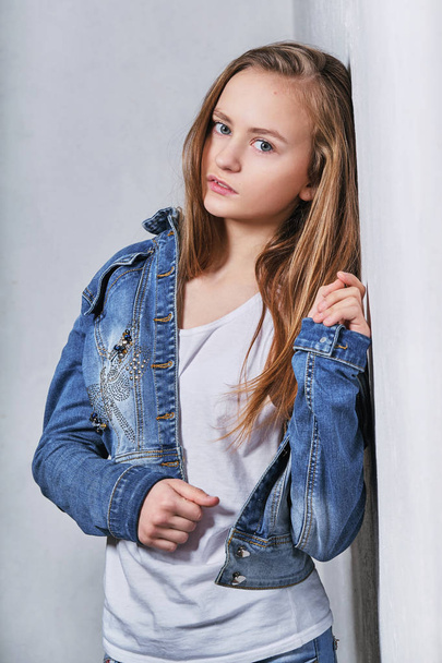 Fashion portrait girl near white wall.Stylish beautiful teenager model clothed in urban casual clothes,jeans jacket. Girl blonde adolescent 11 years posing looking confident,serious face. Model tests. - Foto, Imagem