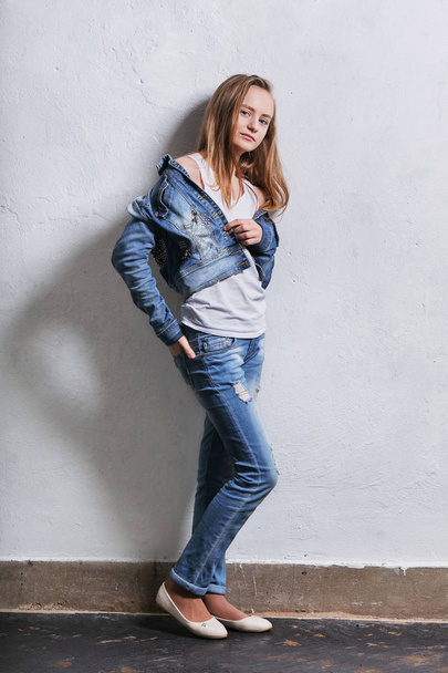 Fashion teenager girl standing leaning back against a white concrete wall. Young caucasian woman 11-14 years in casual stylish clothes, denim jacket, white singlet cotton, youth jeans. - Fotografie, Obrázek