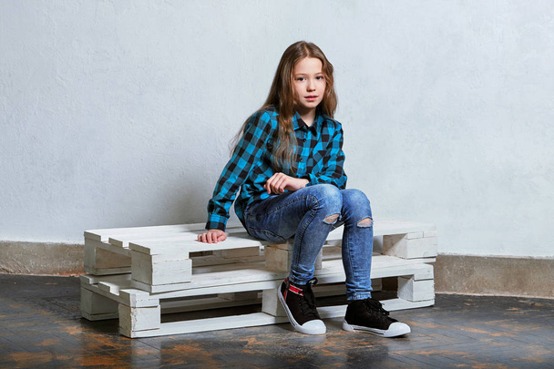 Confident teenager. Stylish serious young girl sitting on white pallet and looking camera. Hipster caucasian child woman in bright blue checkered shirt, jeans, sneakers, casual clothing. - Foto, immagini