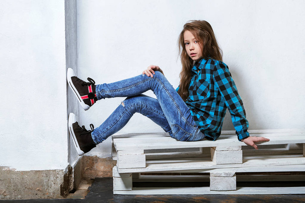 Female teenager sits on pallets near white concrete wall. Girl hipster child in bright blue shirt in a cage,jeans,sneakers.Fashion pose,legs on the wall. Caucasian adolescent in casual clothes. - Foto, Imagem
