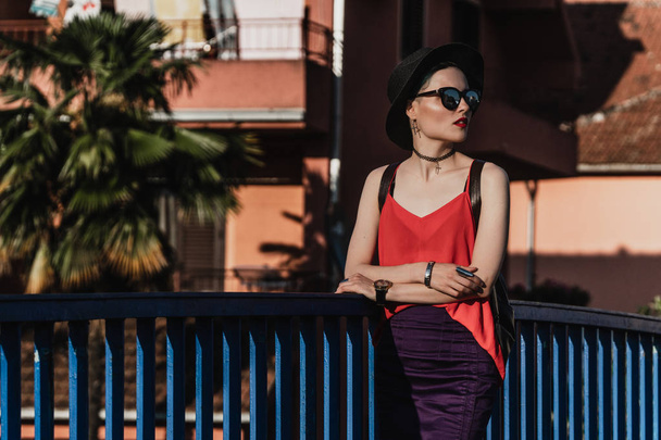 A menina no verão em um chapéu preto e óculos de sol fica na ponte contra o pano de fundo de palmeiras e casas em uma cidade europeia. Viagens e férias
 - Foto, Imagem