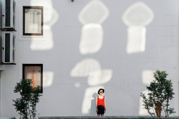Summer girl in a black hat and sunglasses stands near a white wall with reflections from the sun reflected from windows - Valokuva, kuva