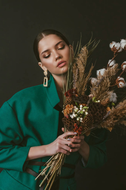 Beautiful girl model with dark hair in a fashionable green jacket with a bouquet of flowers and dried flowers on a black background - Foto, afbeelding