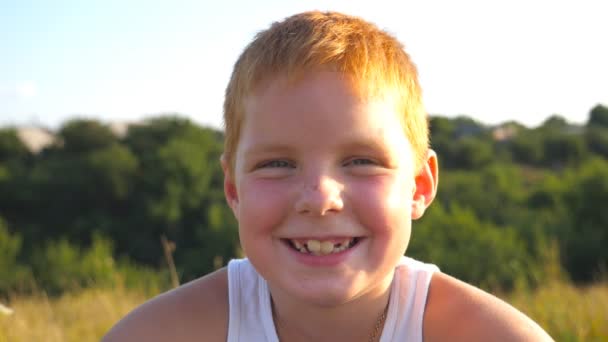 Portret szczęśliwego rudego chłopca z piegami śmieje się na zewnątrz. Urocze, przystojne dziecko patrzące w kamerę z radosnym uśmiechem. Zbliż emocje męskiego dziecka z dobrą miną. Zwolniony ruch - Materiał filmowy, wideo