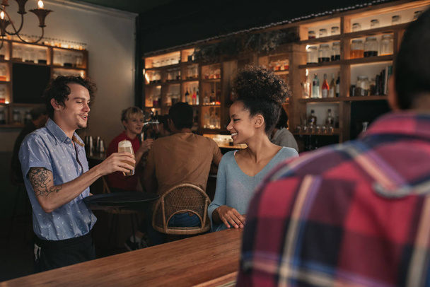 Akşam bir barda birlikte oturan gülümseyen arkadaşlar bir grup için bir içecek sipariş getiren gülümseyen garson - Fotoğraf, Görsel