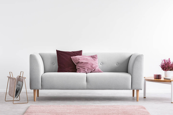 Lilac and burgundy pillows on grey settee in bright living room with heather on coffee table and copy space on the empty white wall - Foto, imagen