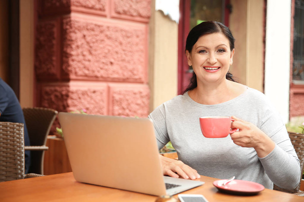 Beautiful mature woman drinking coffee and working on laptop in cafe - Фото, зображення
