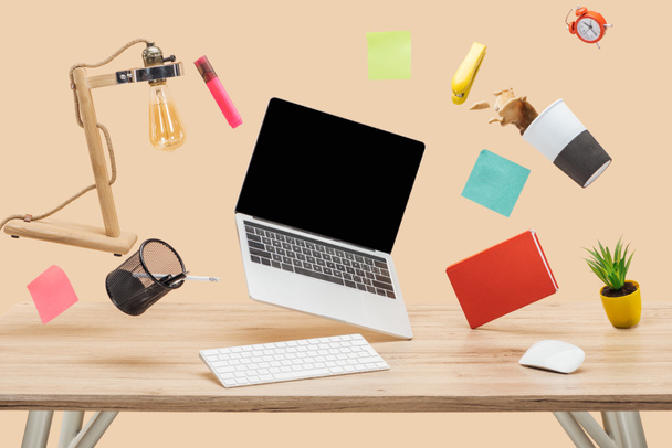 laptop with blank screen, thermomug with coffee splash and stationery levitating in air above wooden desk isolated on beige - Photo, Image