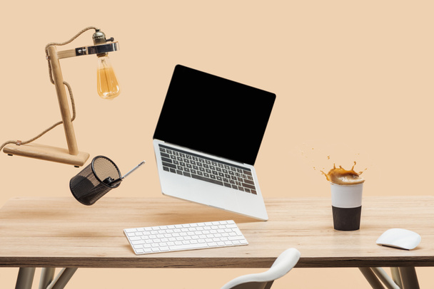laptop with blank screen and lamp levitating in air above wooden desk with thermomug with coffee splash isolated on beige - Photo, Image