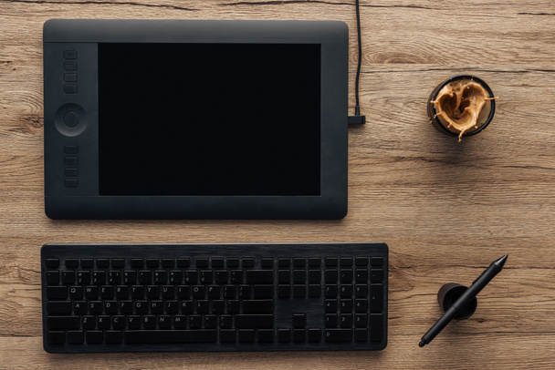 top view of black graphic tablet, graphic pen, wireless computer keyboard and cup with splashing coffee on wooden table - Zdjęcie, obraz