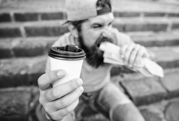 Man bearded shows paper cup drink stairs background. Take coffee with you. Fast food meal for lunch. Hipster bite hot dog hold drink paper cup. Energy from street food daily traditional snack - Фото, зображення