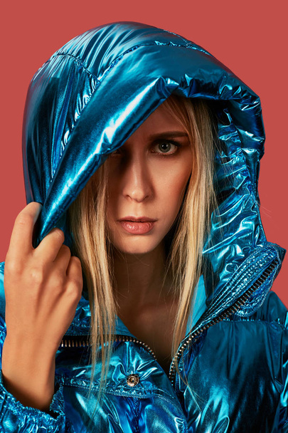 Blonde slim girl in blue down jacket covers her face from wind with hood - Фото, зображення