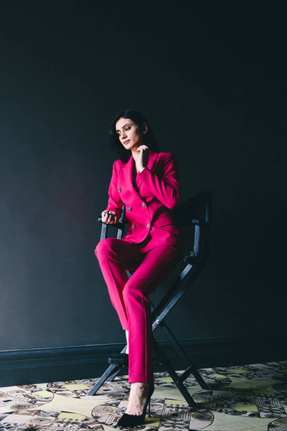 young stylish woman in pink suit posing in dark room with grey walls - Fotó, kép