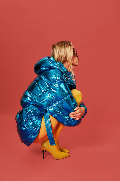 Young cute nice blonde girl in blue jacket sits in a fetal position in studio before camera, red background. Side view - Photo, Image