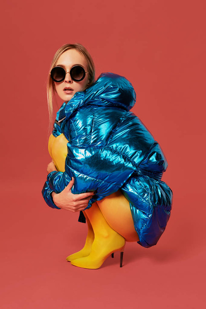 Young cute nice blonde girl in blue jacket sits in a fetal position in studio before camera, red background. Side view - Φωτογραφία, εικόνα