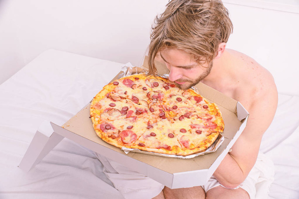 Man likes pizza for breakfast. Bachelors nutrition. Man bearded handsome guy eating cheesy food for breakfast in bed. Guy holds pizza box sit bed in bedroom or hotel room. Food delivery service - Foto, Bild