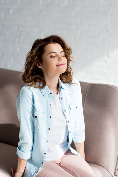 Inspired curly woman in shirt sitting on sofa with closed eyes on grey  - Zdjęcie, obraz