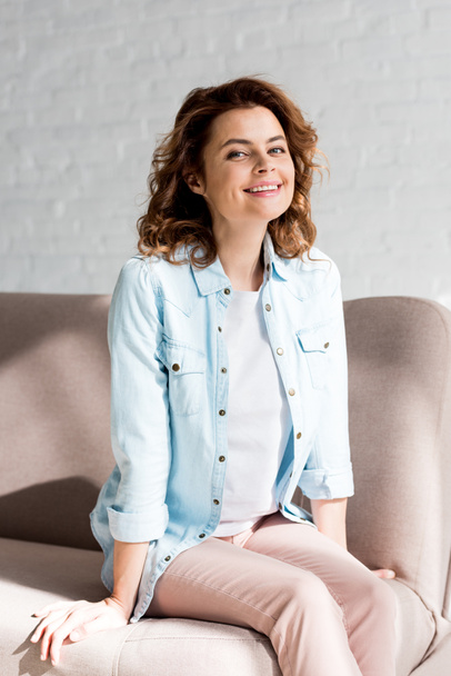 Blissful curly woman in shirt sitting on sofa and smiling on grey  - Φωτογραφία, εικόνα