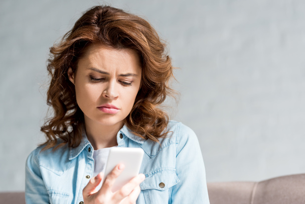 Θλιβερή σγουρά γυναίκα στο πουκάμισο κοιτάζοντας το smartphone στο γκρι  - Φωτογραφία, εικόνα