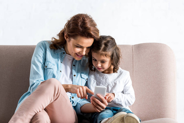 Mother and daughter sitting on sofa and using smartphone on white  - Фото, зображення