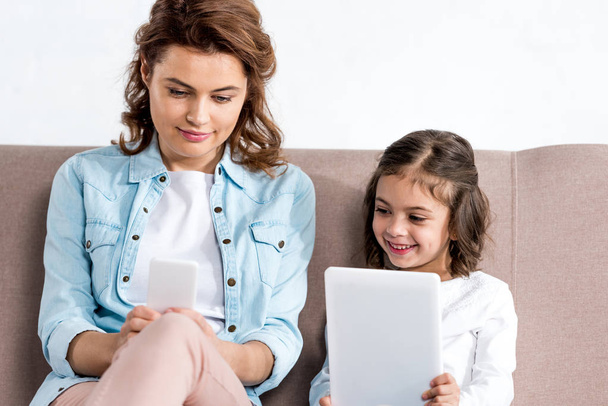 Мати і дочка сидять на дивані і використовують смартфон і цифровий планшет ізольовані на білому
  - Фото, зображення