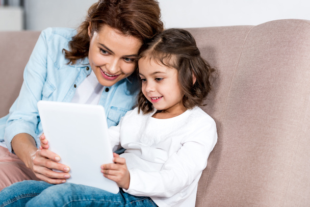 Hymyilevä äiti ja tytär istuu ruskealla sohvalla ja käyttää digitaalista tablettia
  - Valokuva, kuva