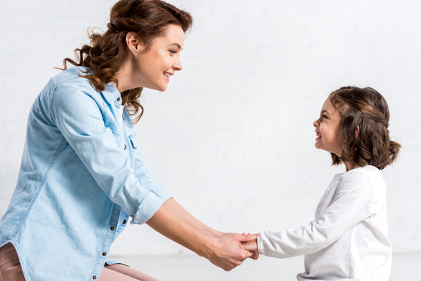 Smiling mother and daughter holding hands and looking at each other on white  - Foto, Imagen