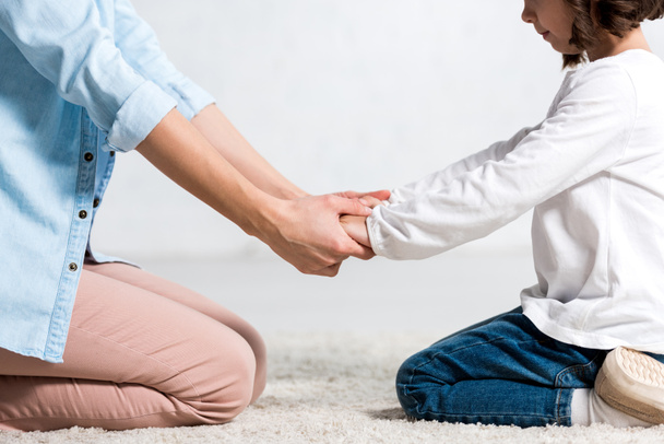 Cropped view of mother and daughter sitting on carpet and holding hands on white  - Valokuva, kuva