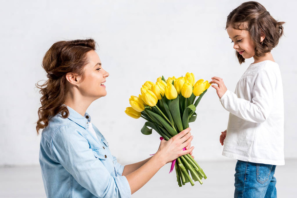 Curly smiling mother and daughter looking at bouquet of yellow tulips on white - Foto, Imagem