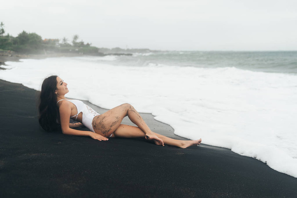 Charming girl with beautiful body lies on black sand beach. Sexy woman rests on summer vacation near ocean. - Фото, изображение