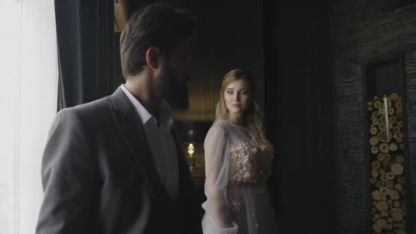 slow motion closeup stern bearded man in elegant suit turns head and looks at attractive blond girl by dark curtain - Filmati, video