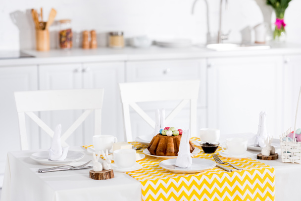 Пасхальный торт с покрашенными яйцами, керамическими кроликами и посуда на столе на кухне
 - Фото, изображение