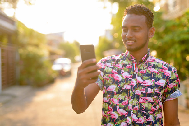 Happy νεαρό όμορφος Αφρικής Τουριστική άνθρωπος χρησιμοποιώντας το τηλέφωνο σε εξωτερικούς χώρους - Φωτογραφία, εικόνα