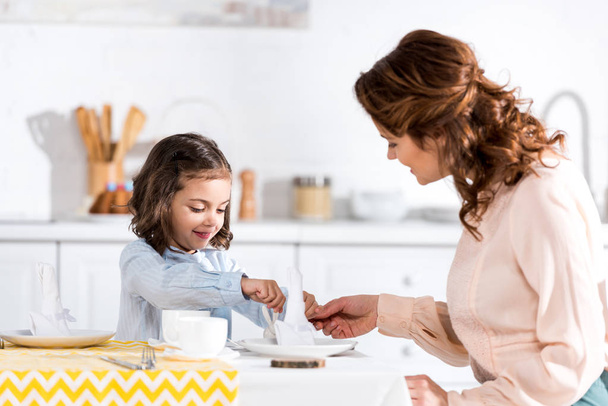 Curly mother and daughter folding napkins at table in kitchen - Photo, Image