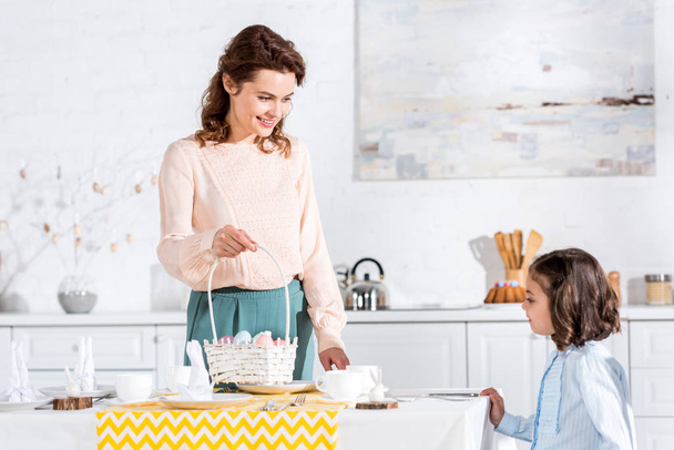 Smiling woman with wicket basket looking at daughter while serving table for easter - Foto, Imagen