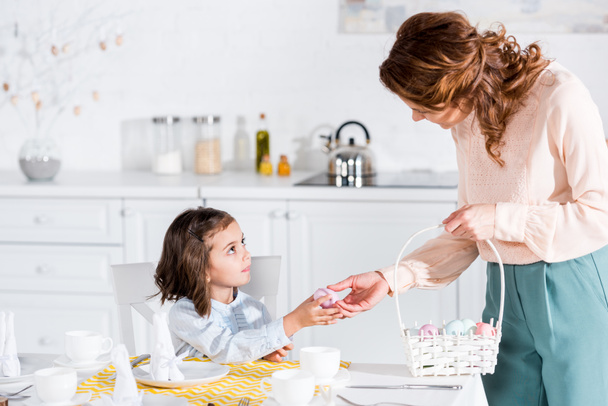 Woman holding wicker basket and giving painted easter egg to daughter in kitchen - Fotoğraf, Görsel