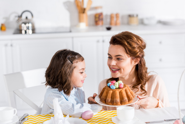Mother showing easter cake with painted eggs to daughter in kitchen - Photo, Image