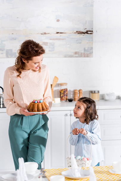 Kid with wicker basket and mother with easter cake looking at each other in kitchen - Photo, Image