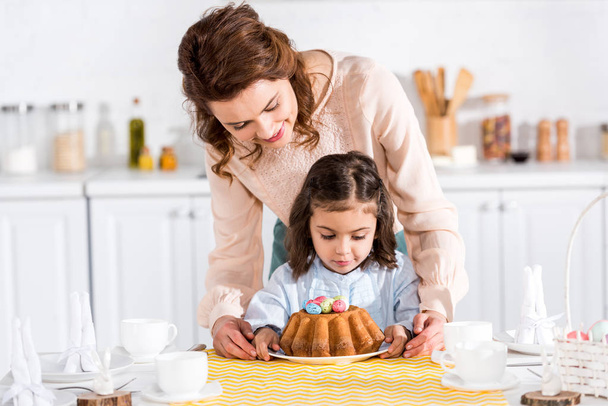 Mother and daughter looking at easter cake with painted eggs in kitchen - Photo, Image