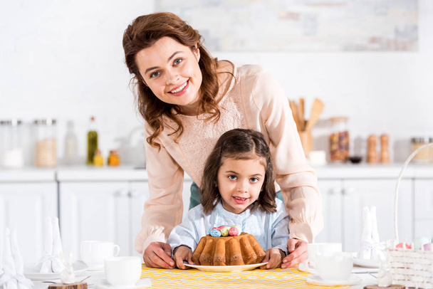 Mãe sorridente e criança com bolo de Páscoa olhando para a câmera na cozinha
 - Foto, Imagem