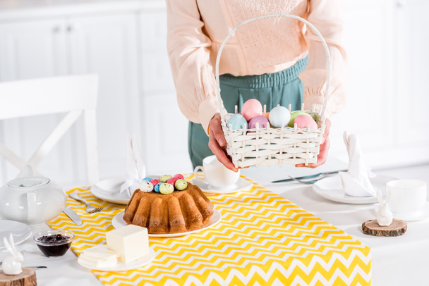 Cropped view of woman holding wicker basket with painted easter eggs near table - Fotografie, Obrázek
