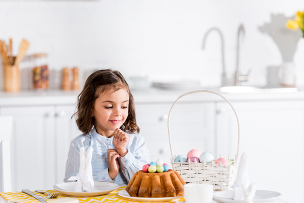 Cute kid sitting at table and looking at easter cake and painted eggs in kitchen  - Fotoğraf, Görsel