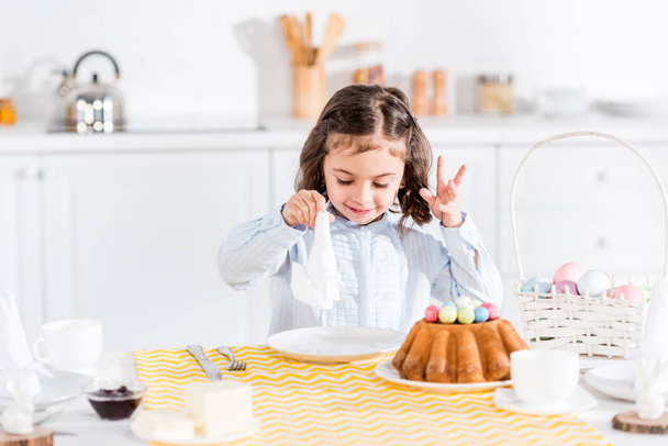 Niño sonriente sentado en la mesa con pastel de pascua y servilleta
 - Foto, Imagen
