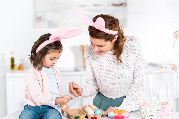 Alegre madre e hija en orejas de conejo pintando huevos de Pascua en la cocina
 - Foto, Imagen