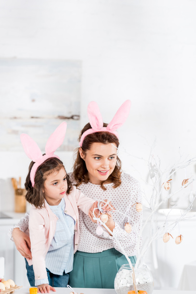 Happy mother and daughter in bunny ears decorating easter tree - Foto, afbeelding