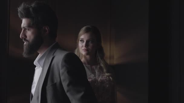 slow motion closeup blonde woman with upset glance stands behind brutal bearded man back in darkness - Imágenes, Vídeo