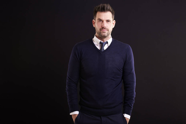 Studio πορτρέτο του αυτοπεποίθηση επιχειρηματίας φορώντας πουκάμισο και γραβάτα με πουλόβερ, ενώ στέκεται με τα χέρια σταυρωμένα στο απομονωμένο μαύρο φόντο. - Φωτογραφία, εικόνα